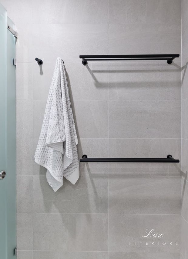 A close-up view of a contemporary bathroom wall finished in large light-grey tiles. Two long matte-black towel rails and a small black hook are mounted on the wall, with a white textured towel hanging from the hook. A frameless or lightly framed glass shower enclosure is visible along the left edge. The space is clean, minimal, and neutral with strong black-and-grey contrast.