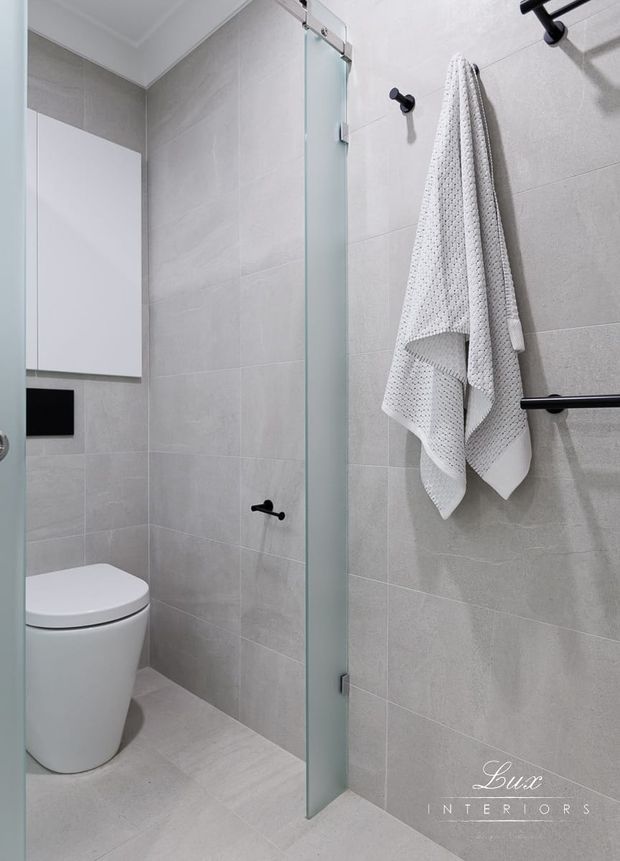 Compact contemporary bathroom with large light-grey tiled walls and floor, a frameless glass shower screen, white wall-mounted storage, streamlined white toilet, and matte black hardware and grab rails. The space has a clean, practical, minimalist appearance.