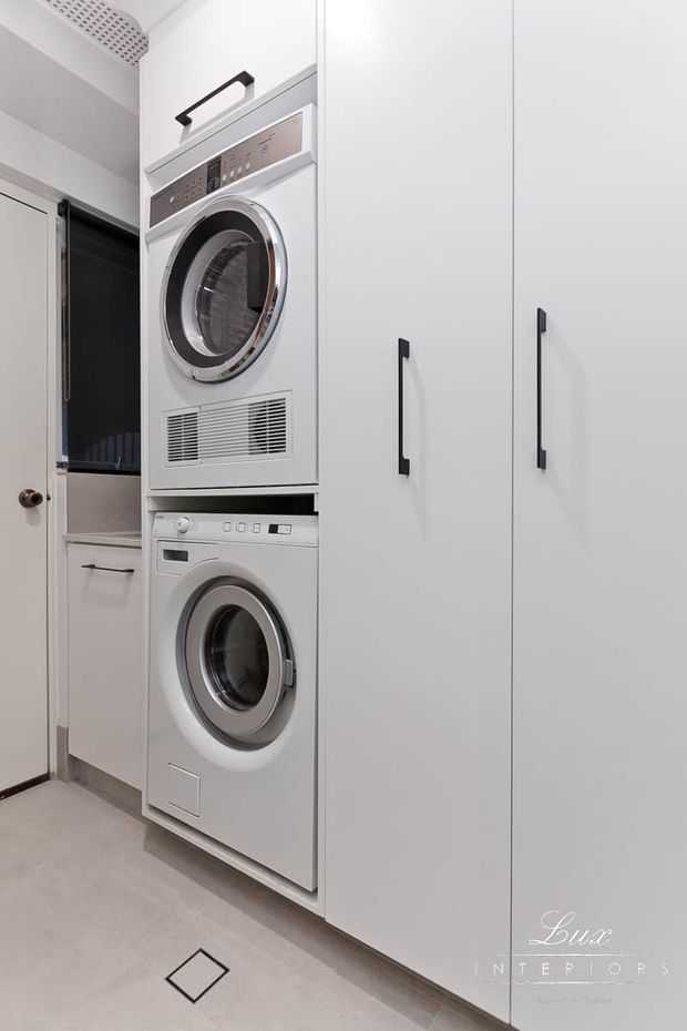 Compact modern laundry room with a vertically stacked front-loading dryer and washing machine integrated beside tall white storage cabinetry. The space features black linear handles, a neutral tiled floor, light walls, and a small utility basin or counter area visible through the doorway.