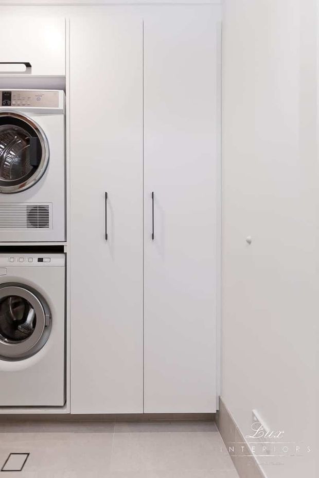 Compact, bright laundry room with a stacked front-loading washing machine and dryer integrated into white cabinetry. The room features tall white storage doors with slim black handles, pale tiled flooring, and a clean minimalist built-in layout.