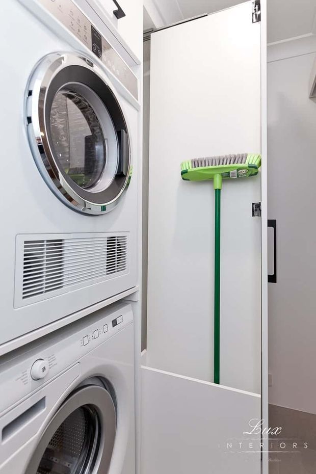Compact laundry room with stacked white front-loading washer and dryer on the left, and a tall white storage cupboard on the right. A green broom is mounted inside the open cupboard door. The space has a clean, practical built-in fit-out with white cabinetry and appliances.