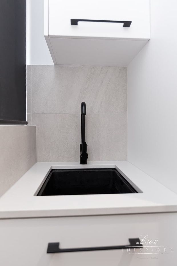 Close-up view of a compact, modern kitchen sink area. A black undermount sink and matching matte-black gooseneck tap are set into a white benchtop, with white cabinetry and black bar handles. The backsplash uses light grey textured tiles, and a dark-framed window is visible at left.