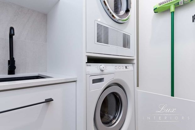 Compact modern laundry room featuring stacked front-loading washer and dryer, white built-in cabinetry, a deep utility sink with a matte black tap, pale tiled walls, and a green-handled cleaning tool mounted on the wall.