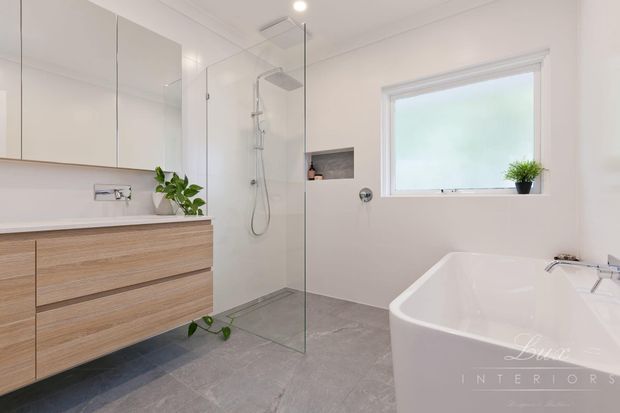 Bright contemporary bathroom featuring a floating timber-look vanity with integrated basin, mirrored wall cabinets, a frameless glass walk-in shower, freestanding white bathtub, grey tiled floor, recessed shower niche, and a frosted window with a small plant.