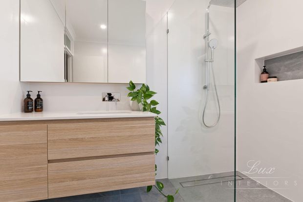 Bright contemporary bathroom with a floating light timber vanity, white countertop and mirrored wall cabinetry. A frameless glass walk-in shower with chrome fittings occupies the right side, complemented by grey floor and shower tiles, a recessed wall niche, and a small indoor plant.