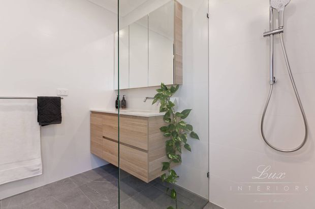 Contemporary bathroom featuring a floating light-wood vanity with integrated drawers, mirrored wall cabinets, a frameless glass shower enclosure, wall-mounted chrome shower fittings, and large grey floor tiles. A trailing plant adds a natural accent to the otherwise clean, light-toned space.