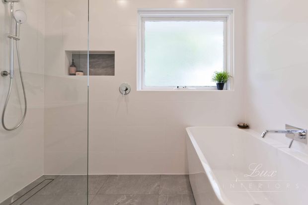 Bright contemporary bathroom featuring a frameless glass walk-in shower, wall-mounted shower fixtures, a recessed shower niche, large-format pale wall tiles, grey floor tiles, a freestanding white bathtub, and a frosted window with a small plant.
