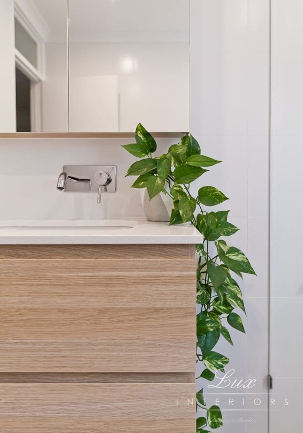Close-up of a refined contemporary bathroom vanity featuring a wall-mounted brushed metal tap, white countertop and backsplash, mirrored cabinetry, and a light timber-grain floating vanity. A trailing green plant adds an organic accent beside the basin.