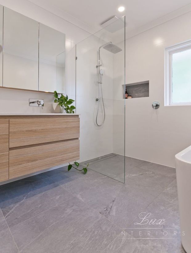 Bright contemporary bathroom featuring a frameless glass walk-in shower with a ceiling-mounted rain shower and handheld fixture, a floating timber-look vanity with integrated basin and mirrored wall cabinets, a recessed shower niche, large grey stone-look floor tiles, and a freestanding bath partially visible at right.