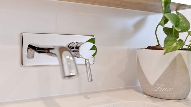 Close-up of a contemporary bathroom wall-mounted chrome tap set with a polished rectangular escutcheon, installed against a pale smooth wall finish. A light stone-look benchtop and a white faceted planter with a trailing plant are visible at the right, with a timber-toned shelf or cabinet edge above.