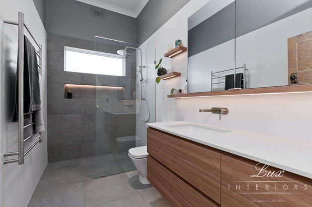 Contemporary bathroom with a walk-in glass shower, wall-mounted rain shower, wall-hung vanity with warm timber cabinetry and white benchtop, large mirrored cabinetry, floating timber shelves, heated towel rail, and grey tiled walls and floor. The room has a clean, refined palette of grey, white, timber, and brushed brass accents.