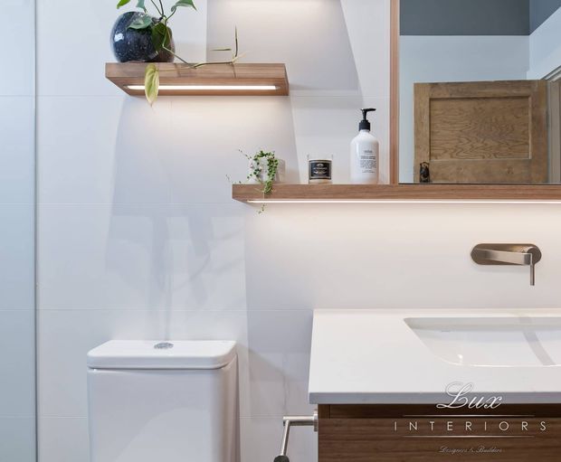 Contemporary bathroom vanity area with a white tiled wall, wall-mounted timber shelves, potted plants, a concealed-cistern toilet, and a white rectangular basin set into a timber-fronted vanity. A large mirror reflects a timber door and part of the room. Brushed metal wall-mounted tapware and warm shelf lighting add refined detail.