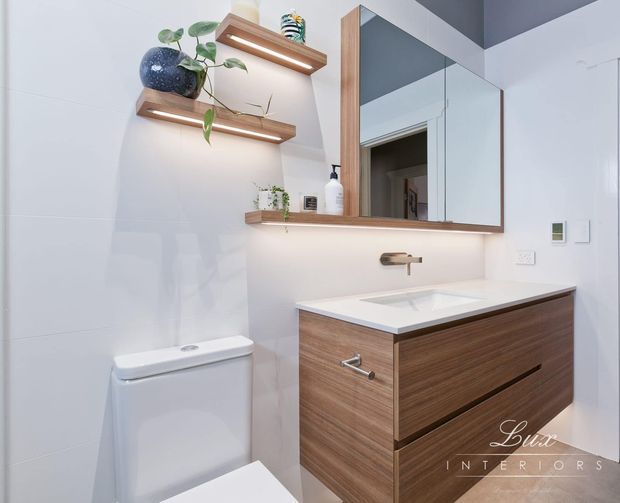 A compact contemporary bathroom featuring a wall-mounted timber vanity with white integrated basin, frameless mirrored cabinet, floating timber shelves with plants and accessories, white tiled walls, wall-mounted tapware, and a concealed-cistern toilet. Warm under-shelf and vanity lighting adds contrast to the clean white finishes.