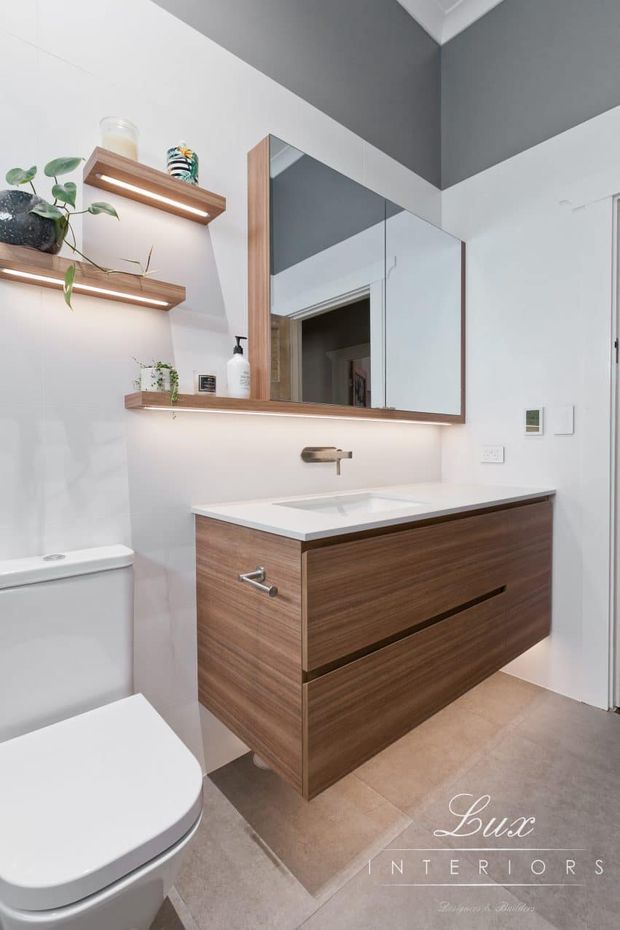 A contemporary bathroom featuring a wall-mounted double-drawer timber vanity with a white integrated basin and matching white benchtop. A large mirrored cabinet, illuminated timber display shelves, concealed wall tapware, white toilet, and warm neutral floor tiles create a clean, refined fit-out.