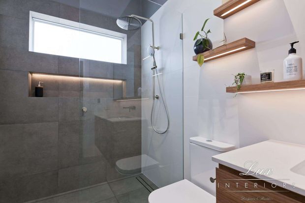 Contemporary bathroom with a walk-in glass shower, large grey wall tiles, recessed and floating timber shelves with integrated lighting, a wall-hung toilet, and a timber-fronted vanity. The space is bright, clean, and modern with indoor plants adding a natural accent.
