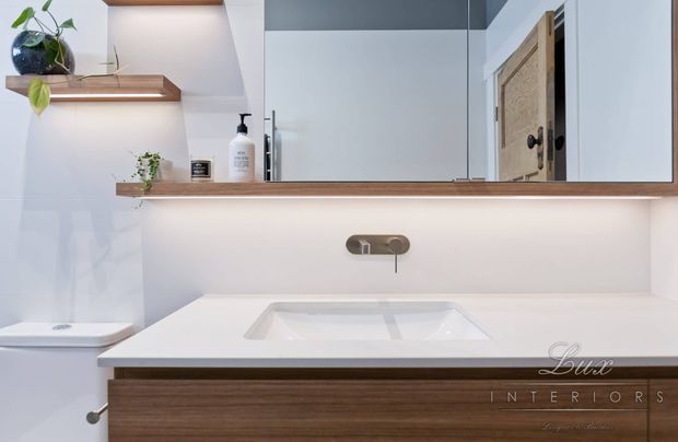 Contemporary bathroom vanity with a white integrated basin and white benchtop, warm timber floating cabinetry, mirrored wall cabinets, brushed metal wall-mounted tapware, and softly lit open timber shelves displaying plants and toiletries. A partially visible toilet is at left.