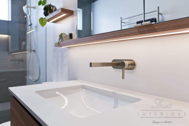 Contemporary bathroom vanity with a white integrated basin, brushed brass wall-mounted tapware, timber drawer cabinetry, and a large mirrored cabinet with warm LED underlighting. A glass-enclosed shower and small built-in timber shelves with plants are visible in the background.