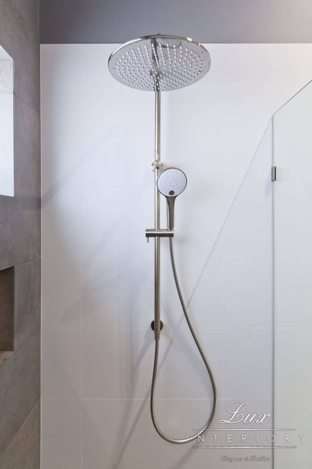 Close-up of a contemporary shower installation with a large round overhead rainfall showerhead, handheld shower, and slim chrome rail mounted against large white wall tiles. A frameless glass shower screen and grey tiled wall are visible at the sides. The image has a clean, minimal bathroom fit-out and includes a Lux Interiors watermark.