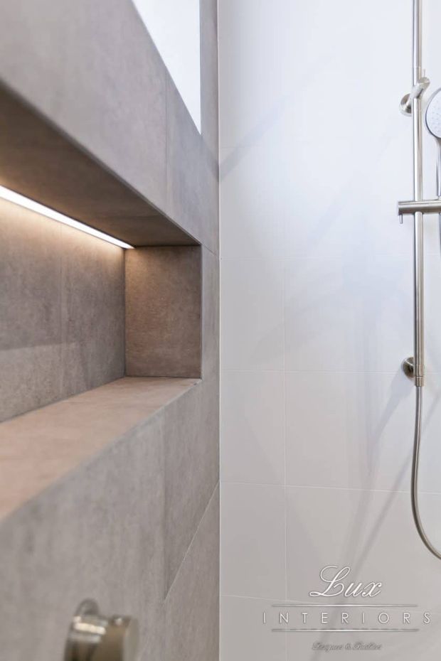 Close-up of a contemporary shower area featuring a built-in recessed niche clad in large-format grey concrete-look tiles, softly illuminated by concealed strip lighting. The adjacent wall is finished in glossy white tile and includes a chrome shower rail and handheld shower fitting.