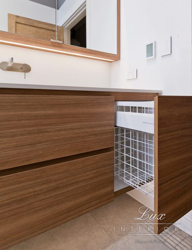 A close-up view of a contemporary bathroom vanity with warm walnut-toned built-in cabinetry, an open wire pull-out laundry hamper, a large framed mirror, and a pale stone or tile benchtop. White walls, integrated lighting beneath the mirror, minimalist wall controls, and a brushed metal wall fixture create a clean, refined fit-out.