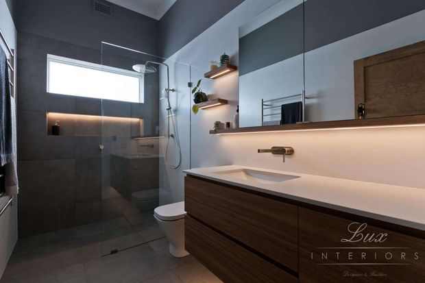 Contemporary bathroom interior with a frameless glass walk-in shower, wall-hung toilet, floating dark timber vanity, integrated white benchtop and basin, large mirror, and warm under-vanity and shelf lighting. The room uses dark grey wall and floor tiles with white geometric wall sections and simple brushed-metal fixtures.