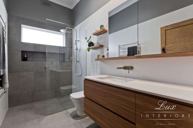 Contemporary bathroom with a walk-in glass shower, wall-hung toilet, large wall mirror, floating timber vanity with a white benchtop, and integrated shelving. The room uses grey tiled walls and floor, white surfaces, warm wood cabinetry, and brushed metal fixtures.