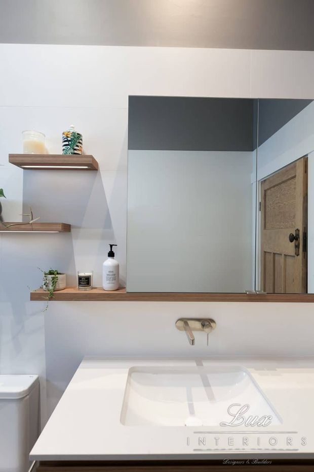 A bright, compact contemporary bathroom vanity area with a large frameless mirror, white tiled walls, a white integrated basin countertop, wall-mounted brushed metal tapware, and warm timber floating shelves and vanity trim. A timber door is reflected in the mirror.