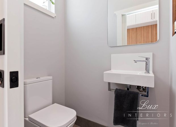 Compact contemporary powder room with a wall-mounted white ceramic basin, chrome tapware, wall-hung plumbing, toilet, and a frameless rounded-corner mirror. Soft grey walls, white fixtures and trim, and dark flooring create a clean, understated scheme. A reflected view shows built-in cabinetry beyond the room.