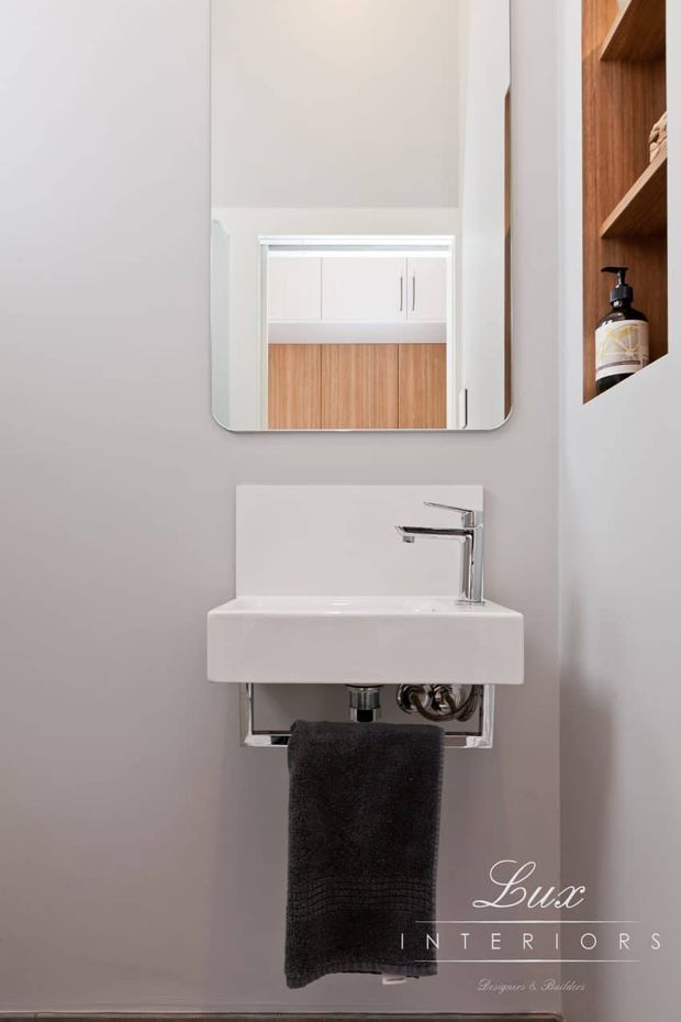 Compact contemporary bathroom or powder room featuring a wall-mounted white rectangular basin with chrome tapware and exposed chrome plumbing support. A rounded-corner frameless mirror hangs above, reflecting white cabinetry and warm timber joinery. Open timber shelving with a soap dispenser is visible at right. The room has light grey walls, a dark towel, and a clean minimalist fit-out.