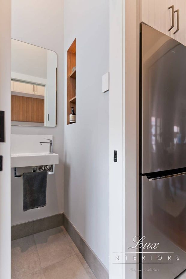 Compact, modern powder room beside a kitchen or utility area, featuring a wall-mounted white basin, chrome tap, rounded-corner mirror, recessed timber shelving, pale painted walls, grey tiled flooring and skirting, and a large stainless-steel refrigerator at the right edge.