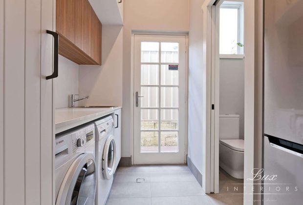 A compact, bright laundry room with white built-in cabinetry, a pale benchtop, timber overhead cabinetry, front-loading washing machines, and a glazed exterior door. A bathroom with a toilet is visible through the open doorway on the right. The space has a clean, contemporary fit-out with light grey floor tiles and white walls.
