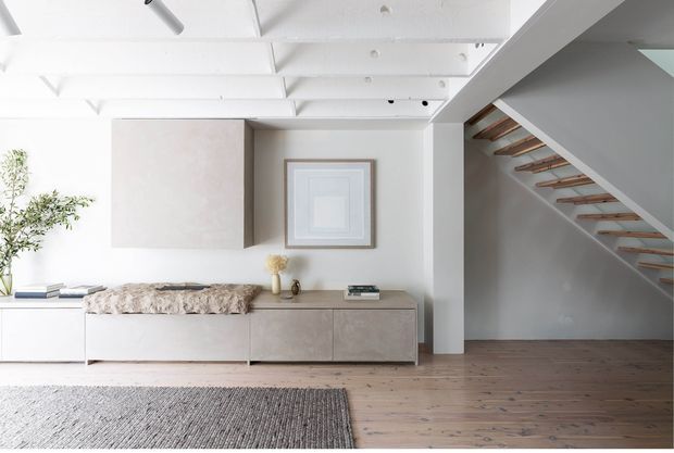 Bright minimalist living area with white walls and exposed white ceiling beams, a low built-in timber-toned media cabinet, framed artwork, soft furnishings, and a timber stair with open risers leading upward. The room has a restrained neutral palette and natural timber flooring.