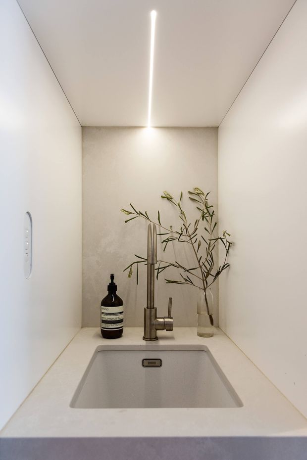 A compact, minimalist sink nook with a deep integrated rectangular basin set into a pale solid-surface counter. The space is enclosed by smooth off-white walls and a ceiling with a slim linear LED light. A brushed metal tap, dark soap dispenser, and a clear vase with leafy branches sit on the counter. A small wall-mounted control panel is visible on the left wall.