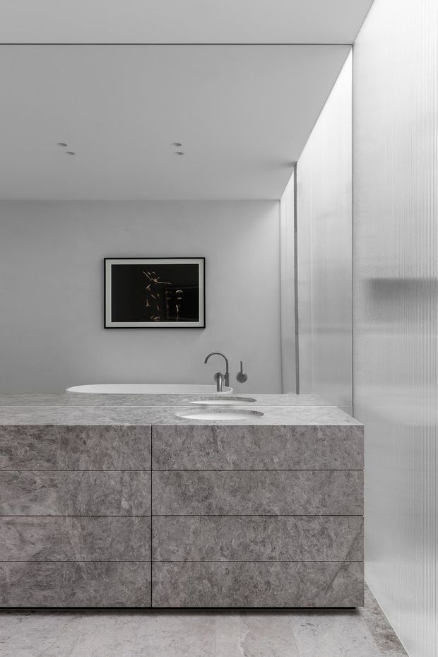 A refined minimalist bathroom featuring a large grey stone vanity with integrated drawers, a pale stone countertop with two basins, a freestanding white bathtub, wall-mounted fixtures, recessed ceiling lights, and a translucent ribbed-glass partition. A dark framed artwork provides contrast against the light neutral surfaces.