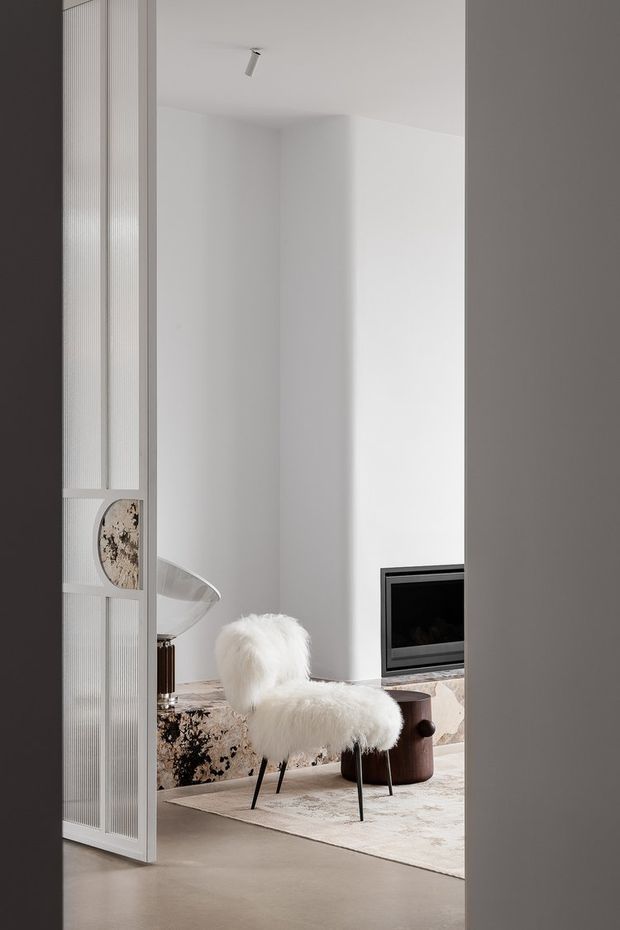 A minimalist living room viewed through a partially open fluted-glass and metal-framed screen door. The bright white space features a sculptural white fireplace wall with a black inset fireplace, a white sheepskin lounge chair, a dark wood side table, a patterned low sofa or bench, and a pale area rug. Neutral finishes and soft natural light create a refined, serene atmosphere.