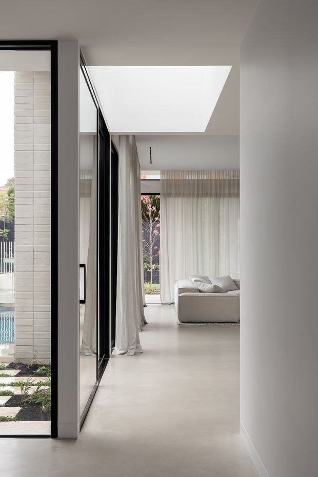A minimalist residential hallway opening into a bright living room, with polished pale flooring, white walls, full-height sheer curtains, black-framed internal glazing, and a large angular skylight or clerestory opening. A neutral sectional sofa is visible in the living area, with a glimpse of landscaped exterior and pool through the glazing.