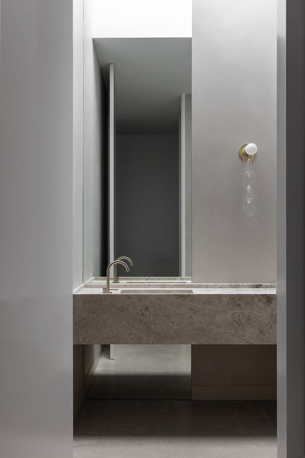 A minimalist bathroom vanity viewed through a doorway, featuring a thick grey natural-stone benchtop with integrated basin and brushed brass curved tapware. A large frameless mirror, grey cabinetry, pale grey walls, and understated pendant-style lighting create a refined monochromatic interior.