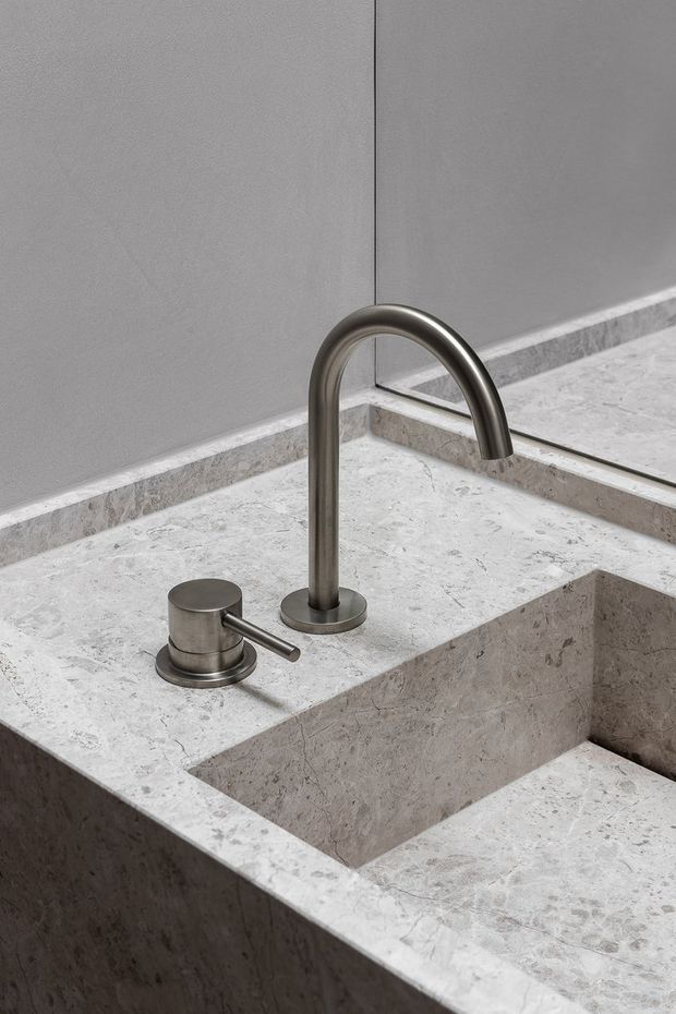 Close-up of a minimalist bathroom vanity featuring a monolithic pale grey natural-stone benchtop and integrated rectangular basin. A brushed metal gooseneck mixer tap and separate cylindrical control are mounted beside the basin, with a large mirror and matching stone splashback detail behind.