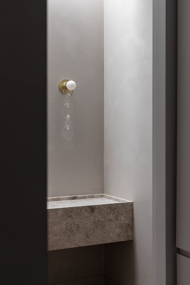 A narrow, minimalist interior alcove featuring a wall-mounted brass-and-white spherical fixture above a thick stone ledge or bench. The space has smooth pale walls, dark cabinetry or framing at the sides, and a warm, subdued palette with strong vertical lines.