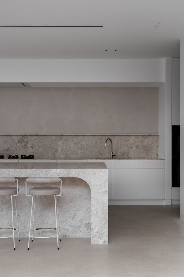 A minimalist contemporary kitchen with a pale stone island featuring a rounded waterfall end and integrated seating. The kitchen includes white handleless cabinetry, a textured stone backsplash, a slim dark cooktop, and a curved brass-toned tap. Neutral plaster, stone, and concrete-like finishes create a calm, refined aesthetic.