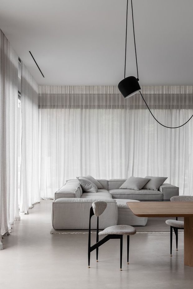 A bright, minimalist open-plan living and dining area with floor-to-ceiling sheer white curtains, a large pale grey sectional sofa, a timber dining table, and sculptural upholstered dining chairs. A black pendant light with a long curved cord provides contrast against the white ceiling and soft neutral interior.