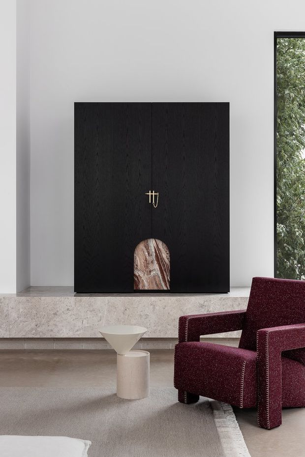 A refined contemporary living room featuring a minimalist black timber fireplace surround with a small marble or stone inset, set above a pale stone hearth. A burgundy upholstered armchair and sculptural side table sit on a neutral rug, with a narrow black-framed window revealing greenery outside. The palette is restrained and sophisticated, combining dark timber, pale stone, warm grey flooring, and deep red upholstery.