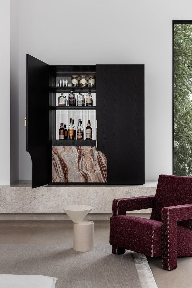 A refined contemporary living room featuring a tall, dark timber built-in bar cabinet with open shelves displaying bottles and glassware. The cabinet incorporates a dramatic veined stone panel, set against a pale wall above a broad stone-clad ledge. A deep burgundy upholstered armchair, sculptural side table, and neutral rug complete the sophisticated interior.