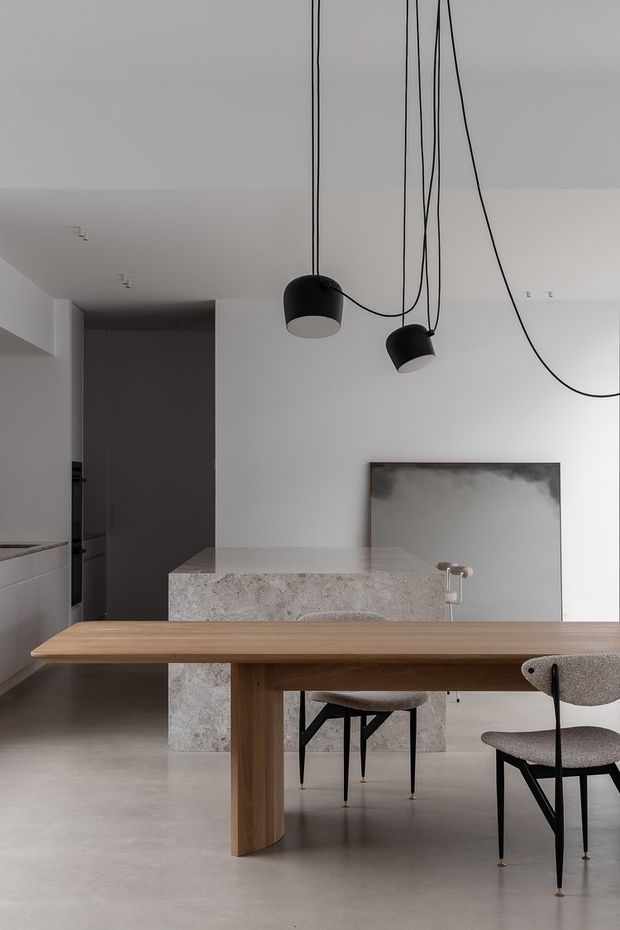 A minimalist open-plan dining and kitchen interior with a long light timber dining table, sculptural upholstered chairs, a pale stone kitchen island, and a monochromatic grey-white palette. Black pendant lights hang from long exposed cords above the table, while a large abstract artwork leans against the wall.