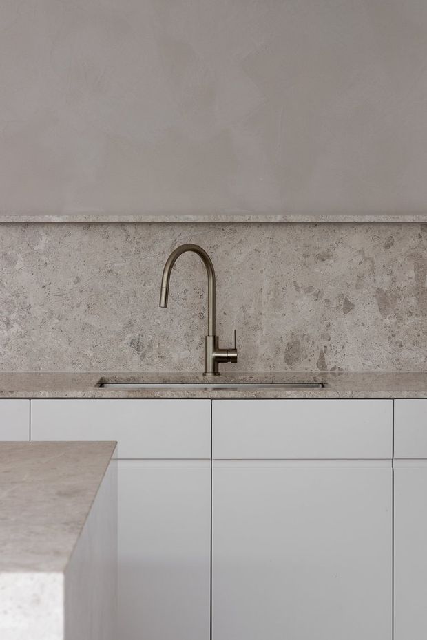 Close-up of a refined minimalist kitchen, showing a pale stone benchtop and integrated sink beneath a tall aged-bronze gooseneck tap. The backsplash is clad in mottled natural stone, with smooth light cabinetry and a partial island in the foreground. The palette is soft grey, beige and off-white with understated, warm detailing.