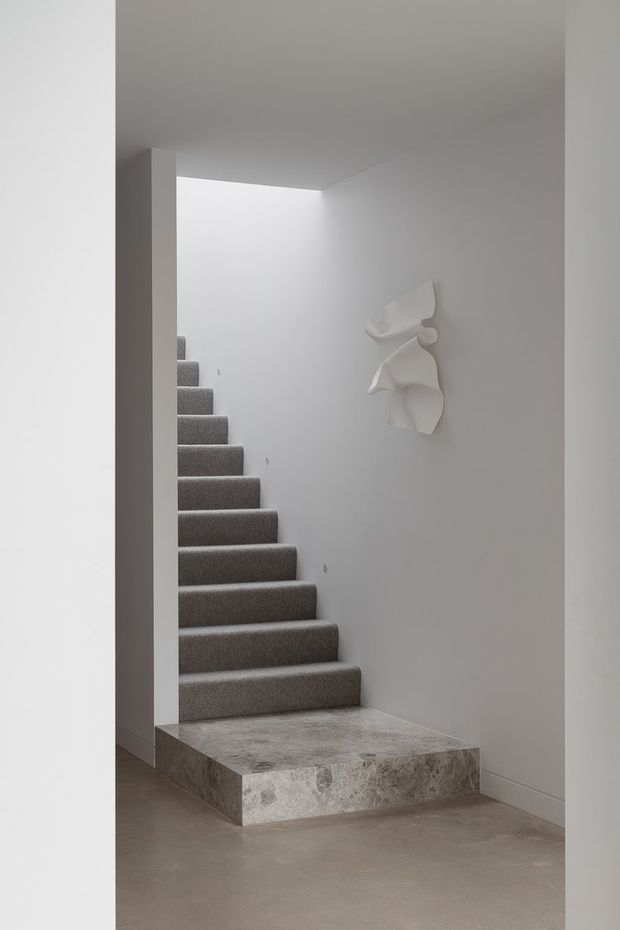 Minimalist interior view of a stairway ascending from a raised stone-clad landing. The stairs are fully carpeted in medium grey, with smooth white walls, a plain white ceiling, and a sculptural white wall artwork. The lower floor appears to be smooth grey concrete or stone, creating a restrained monochromatic palette.