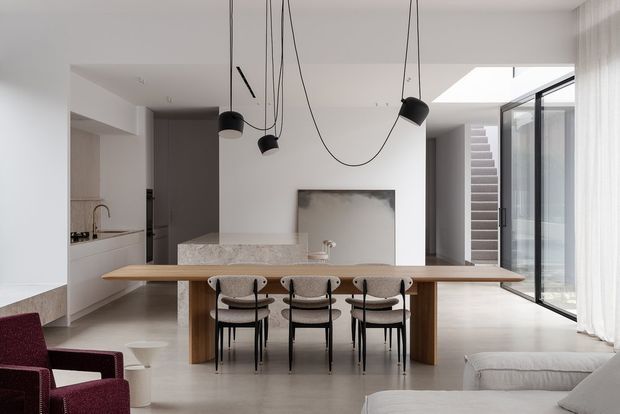 Open-plan contemporary dining and living area with a long timber dining table and four upholstered chairs, adjoining a minimalist white kitchen. Sculptural black pendant lights hang over the table, while full-height glazing and a skylight illuminate the neutral interior. A stairway and outdoor area are visible beyond the glazing.
