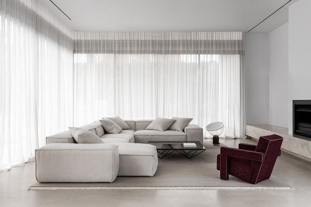 A bright, minimalist living room with floor-to-ceiling sheer curtains, a large pale modular sectional sofa, a low dark coffee table, burgundy lounge chairs, and a sleek linear fireplace set into a pale stone surround. The space has a refined neutral palette, clean-lined built-in detailing, and a contemporary open feel.