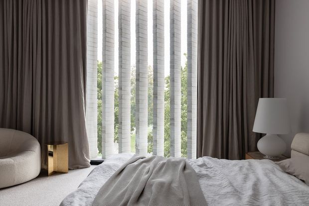 A refined bedroom interior with a low upholstered bed, softly rumpled white bedding, a rounded lounge chair, and a sculptural bedside lamp. Full-height dark taupe curtains frame tall narrow windows with translucent vertical screening and views of greenery, creating a calm, softly lit atmosphere.