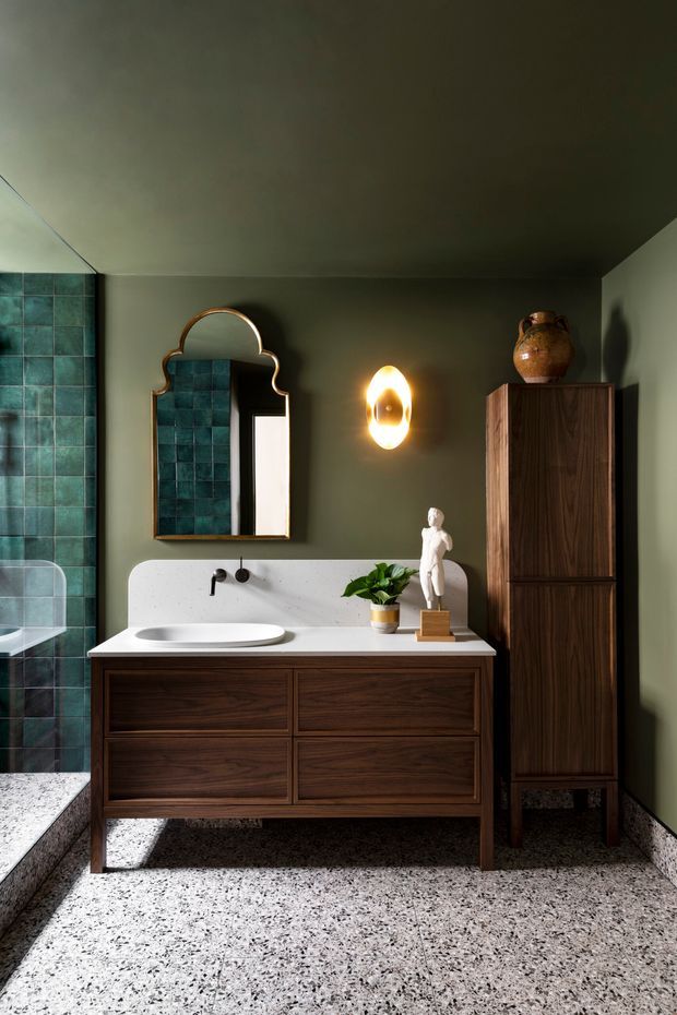 A richly styled bathroom featuring a dark timber double vanity with white integrated basin, green painted walls, teal-green wall tiles in the shower area, terrazzo flooring, an ornate arched mirror, sculptural wall lighting, and a tall timber storage cabinet. The space has a moody, refined, mid-century-inspired character with decorative objects and greenery.
